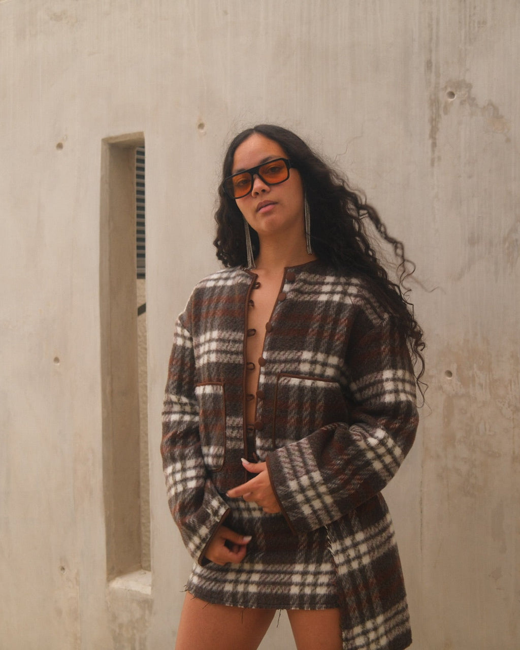 Woman wearing a plaid outfit standing against a concrete wall.