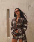 Woman wearing a plaid outfit standing against a concrete wall.
