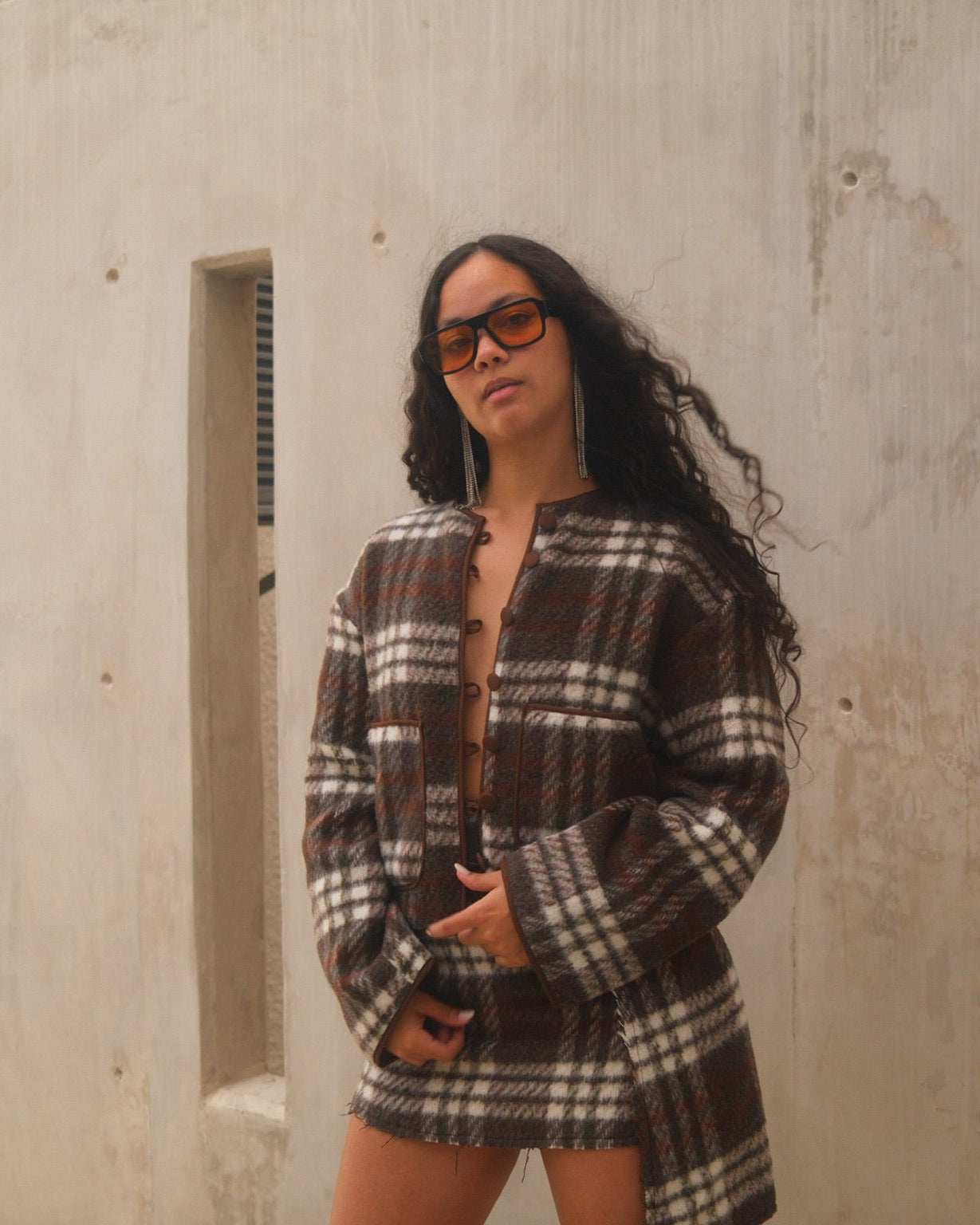 Woman wearing a plaid outfit standing against a concrete wall.